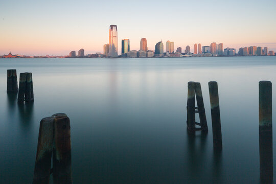 Jersey City skyline and wooden harbor pilings captured at sunset over calm waters, representing a serene tourism destination with atmospheric copy space, Jersey City, New Jersey, USA.