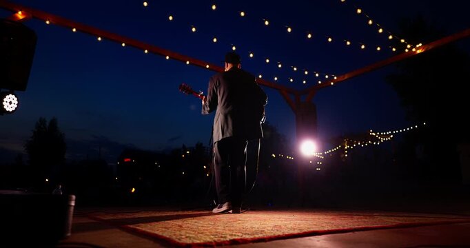Silhouetted guitarist on outdoor wooden stage at dusk under string lights