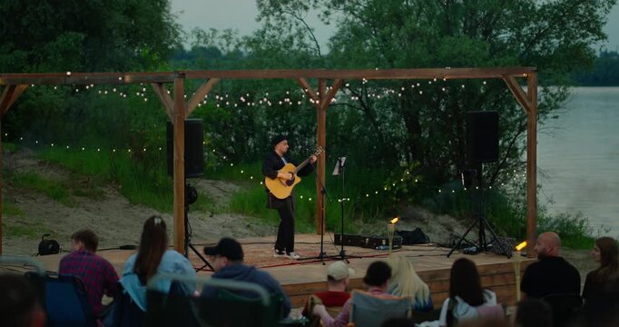 Male guitarist performing on a small lakeside wooden stage at dusk