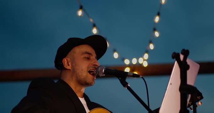Male singer playing acoustic guitar on dim outdoor stage at dusk