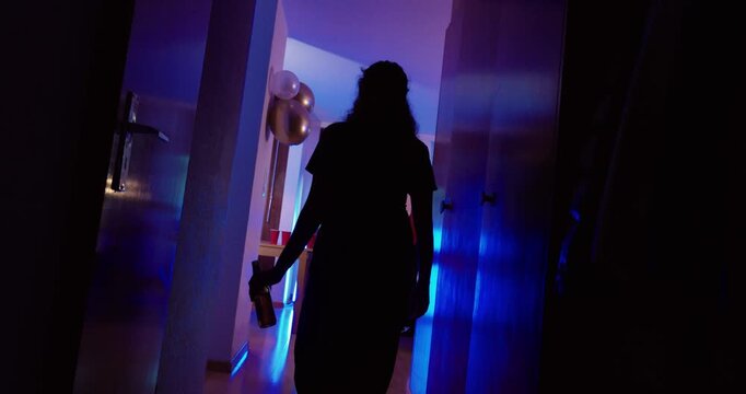 Atmospheric stock video showing a young person holding an alcohol bottle while entering a party environment with dark lighting. Suitable for youth drinking, alcohol consumption, nightlife, peer pressu