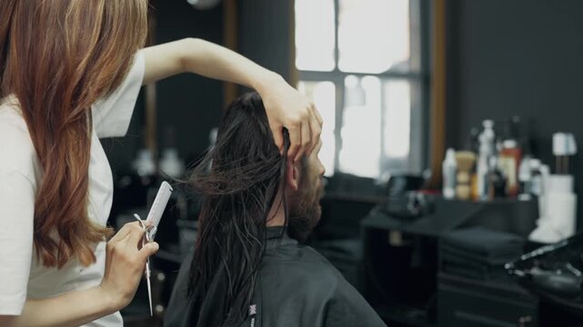 Female barber cutting long wet hair of male client with scissors and comb during professional haircut procedure in modern barbershop