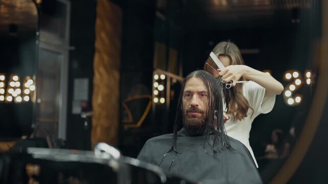 Female barber cutting long wet hair of male client with scissors and comb during professional haircut procedure in modern barbershop