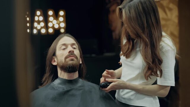 Professional female barber trimming beard of male client with electric clipper during grooming procedure in modern barbershop