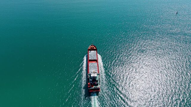 Following a small general cargo vessel sailing across calm turquoise coastal waters. Modern freighter from drone footage.