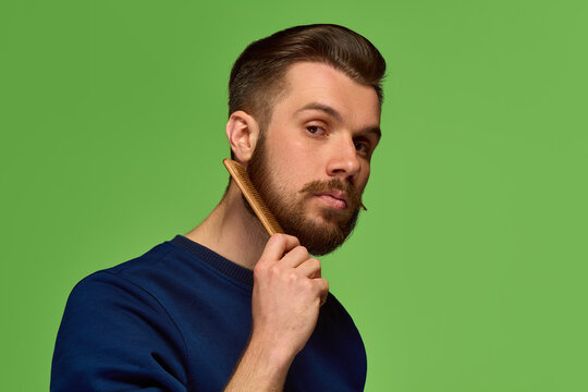 Bearded man holding wooden comb near face on green background. Concept of beard grooming, male beauty care, barbershop branding and personal style advertising.