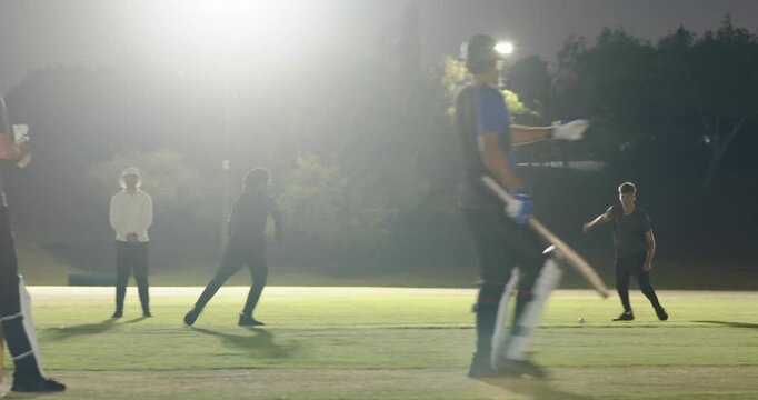 Teenage boy bowling batsman driving offside players retrieving returning under floodlights