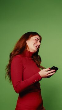 Happy woman in red holds her smartphone against green screen background. She laughs, dances, and enjoys her time. Perfect for advertising.