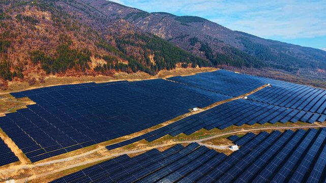 Photovoltaic power station at the base of forested mountain slope. Dense panel rows fill the frame with autumn trees and rocky terrain above.