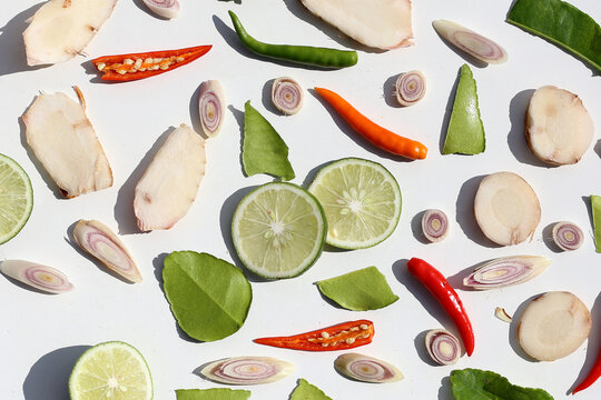 A colorful, close-up pattern of Thai herbs: lime slices, galangal, lemongrass, chilies, and kaffir lime leaves.