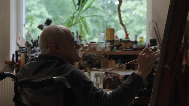 Senior artist on a wheelchair paints an oil painting on canvas in a studio