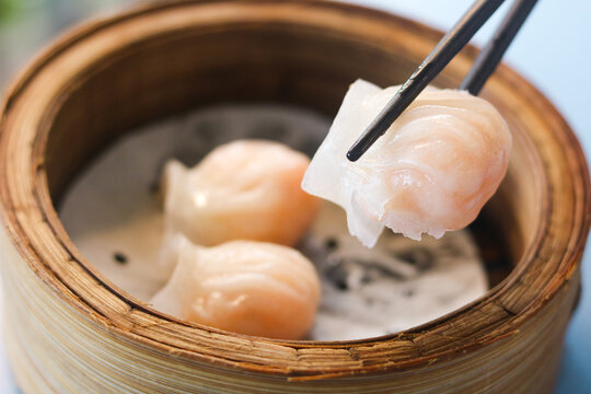 Eating steamed har gow dumplings with chopsticks. It is served in a bamboo basket with translucent wrappers showing the shrimp filling inside.