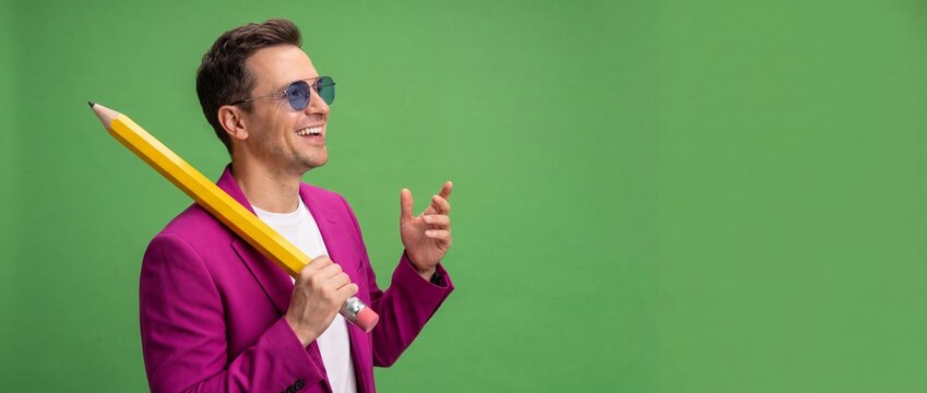 Man holding giant yellow pencil in magenta blazer posing on solid green background chroma key with banner copy space, representing creative writers, artist inspiration, humour, and fun school concepts