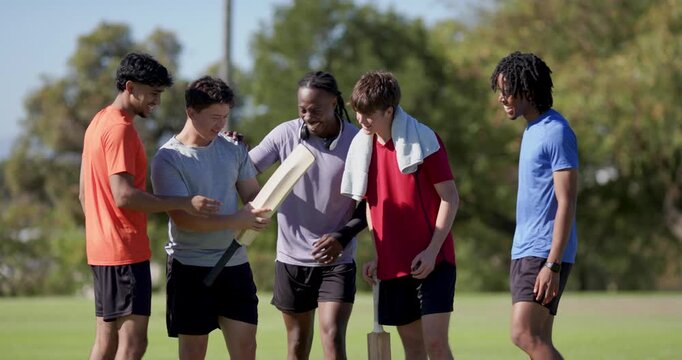 Diverse male teammates on pitch examining bat stacking hands cheering preparing match