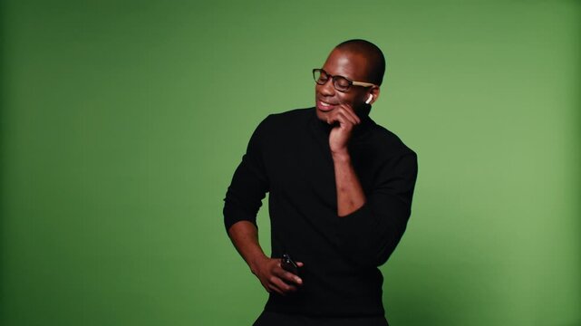 A stylish African-American man dances and enjoys music on his phone against a vibrant green screen background, creating a dynamic and cheerful visual.