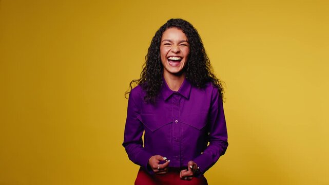 Young woman with curly hair laughs heartily against a vibrant yellow backdrop. Her genuine joy and positive energy fill the frame, creating a cheerful and uplifting scene.