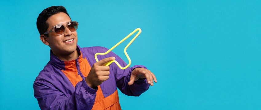 Smiling man holding neon sign arrow pointing up, wearing retro vintage windbreaker jacket and sunglasses, posing against blue studio background banner with copy space for advertising. Cool 90s style..
