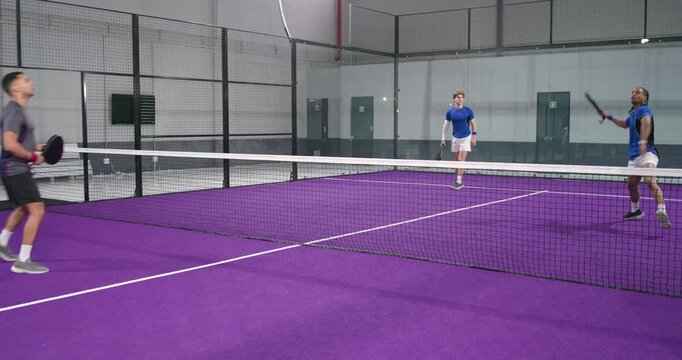 Diverse male teams initiating serve, exchanging racket volleys to score point on purple padel court