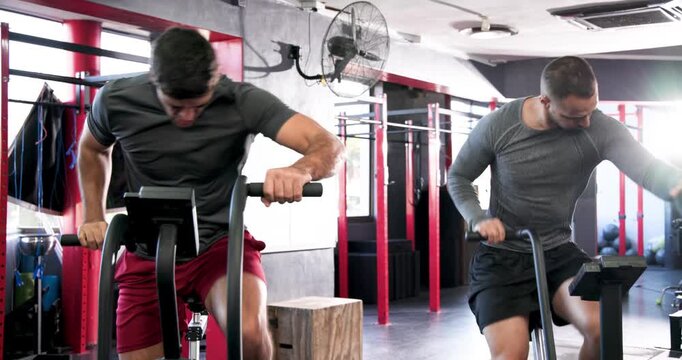 Diverse male friends mounting adjacent air bikes at gym, pedaling hard