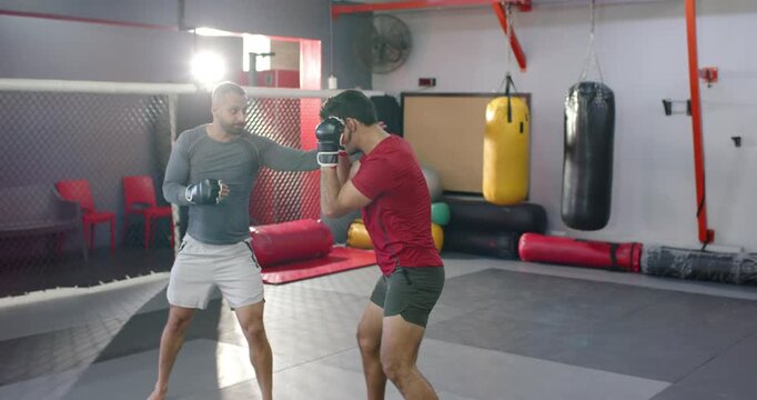 Diverse male fighters with trainer raising gloved hands in gym, trainee practicing blocks