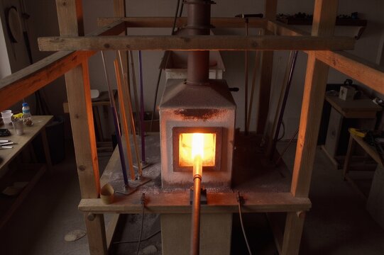 A small refractory kiln, its dark brown exterior glowing warmly in the fluorescent-lit glassblowing workshop, heats a copper pipe to a vibrant orange-yellow as it stretches from