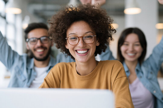 Bright smiles fill a modern office space. Curly-haired woman beams at the camera from her laptop. Behind her, colleagues cheer with raised arms. Warm tones glow under soft ceiling lights