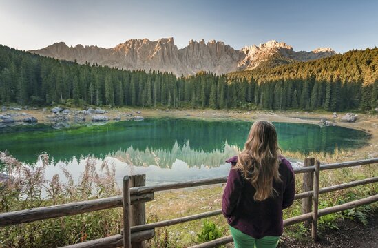 Woman overlooks Lake Carezza, Lago di Carezza, morning sun, Latemar group reflected in the lake, mountain tops Torre Diamantidi, Eastern Latemar peak, Col Coron, Catfish, Bolzano, South Tyrol, Italy