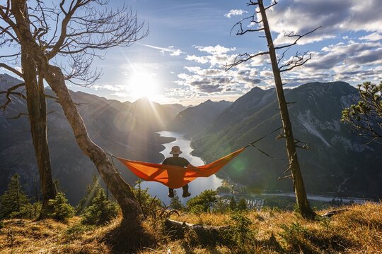 Man with a sun hat sitting in an orange hammock, panoramic view of mountains with lake, Plansee, Tyrol, Austria