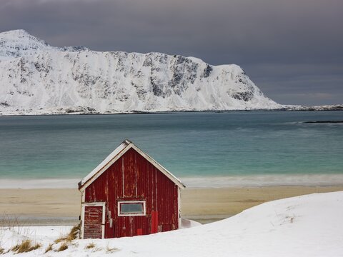 Red Rorbuer fisherman's hut on the beach in the snow, snow-covered mountains behind, Ramberg, Flakstad&oslash;ya, Lofoten, Norway