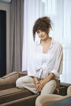 A young woman with curly hair sits comfortably on a couch, wearing a white blouse and beige pants
