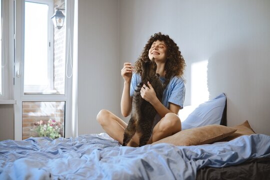 A woman sits on her bed with pillows and blue sheets. She smiles while holding and playing with a black cat. Bright light comes in through a window, creating a warm atmosphere