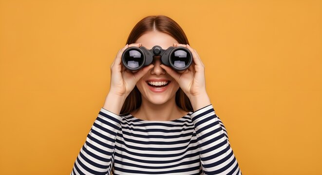 Cheerful young woman smiling while holding binoculars, looking forward with curiosity against a vibrant studio background
