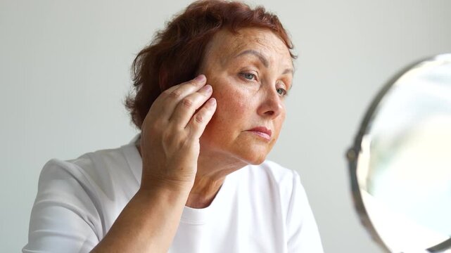Adult woman examines wrinkles in a small mirror indoors. An older woman looks at reflection in front of mirror and touches her face with her hands. Concepts of beauty, care, and aging.