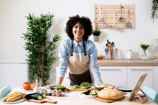 Portrait of young woman leaning on kitchen table and smiling at camera
