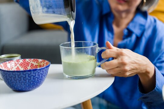 Woman pouring milk into a glass of matcha tea at home