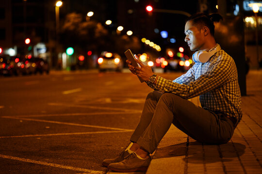 Young man sitting at roadside in the city at night using tablet