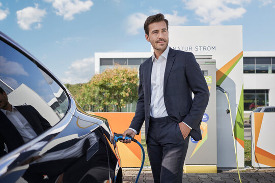 Smiling young businessman holding plug by car at electric vehicle charging station