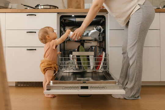 Baby helping mother load dishwasher in home kitchen