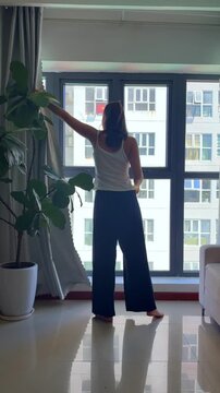 Close-up video of a woman pulling back window curtains to let natural light into a room. Lifestyle scene representing morning routine, home comfort, fresh start and everyday living.