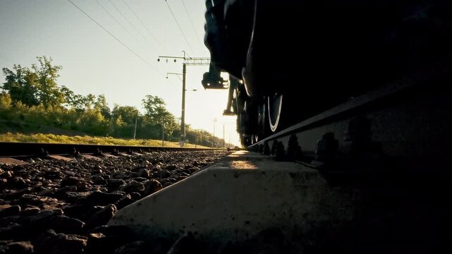 Close-up of a freight train's wheelsets rolling over iron rails across a sun-lit countryside. The scene highlights smooth railway transportation, emphasizing both freight and passenger movement