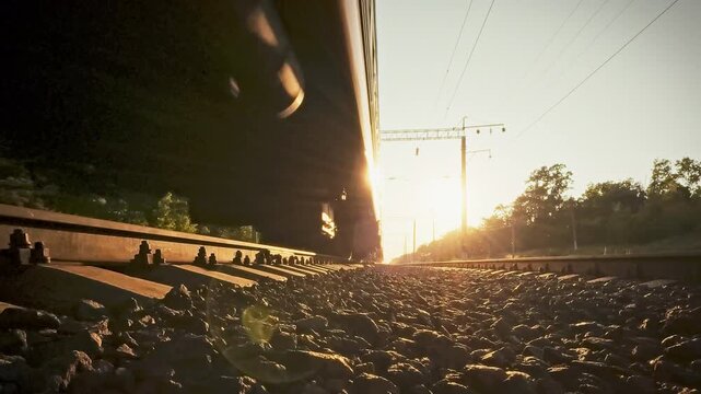 Close-up of passenger train's wheels rolling over iron rails across sun-lit rural landscape, afternoon glow highlighting motion. It illustrates vital rail transportation linking remote communities