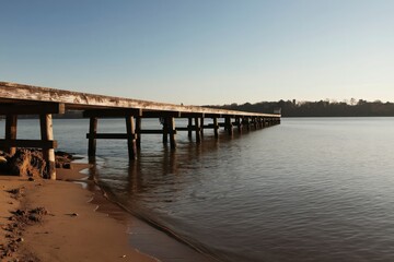 Obraz premium Long wooden pier extends over calm water. Sandy shore meets still lake on clear winter day. Bare trees line distant horizon under pale blue sky.