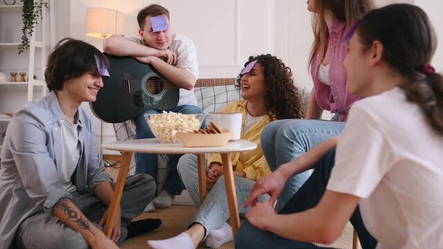 Young adults engaged in a guessing game with cards on their foreheads, seated on the floor in a cozy living room, surrounded by snacks and a guitar
