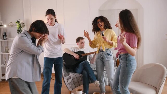 Young adults celebrate in a stylish living room, one playing guitar while others dance and sing, with snacks and drinks on a coffee table