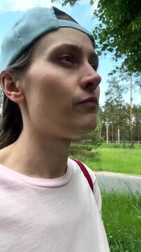 A young woman wearing a baseball cap backwards walks on the street near a pine forest