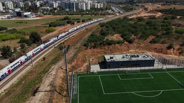 4K aerial drone footage tracking passenger train departing Rehovot toward Beer Yaakov, passing beside the new football stadium with urban landscape, transportation infrastructure and motion cinematic