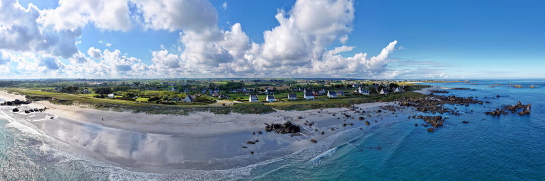 Ein weites Panorama vom Plage de Boutrouilles mit hellem Sand, t&uuml;rkisblauem Meer, den typischen Granitfelsen und einer l&auml;ndlichen Siedlung unter markantem Wolkenhimmel.