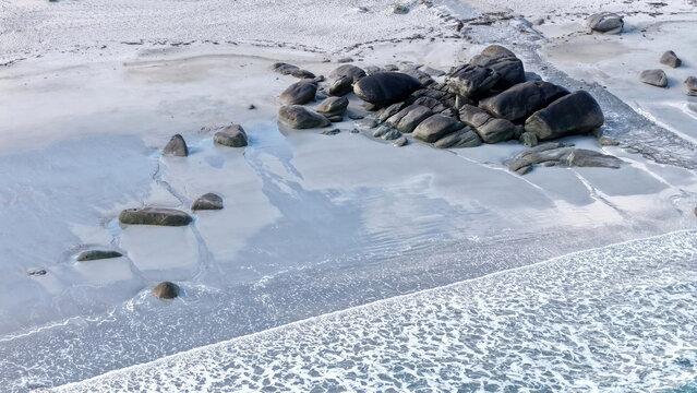 Eine beeindruckende Luftansicht vom Plage de Boutrouilles, bei der sanfte, wei&szlig;e Wellenmuster auf den hellen Sandstrand und die charakteristischen, dunklen Felsformationen treffen.