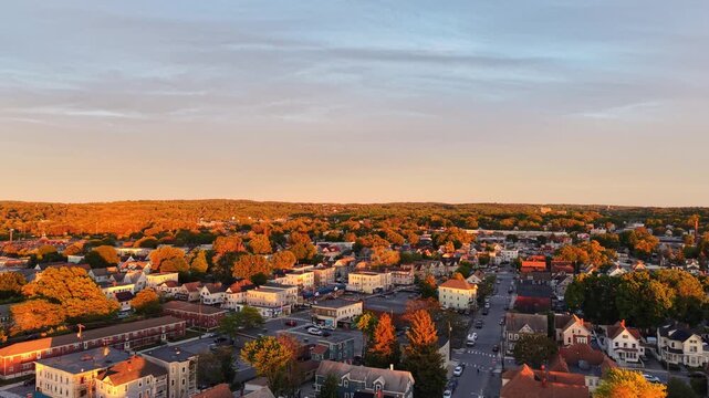 Dense residential neighborhood with mixed rooftops and golden autumn tree canopy glowing under warm sunset light stretching toward forested horizon in Lawrence, Massachusetts, USA.Aerial drone flyover