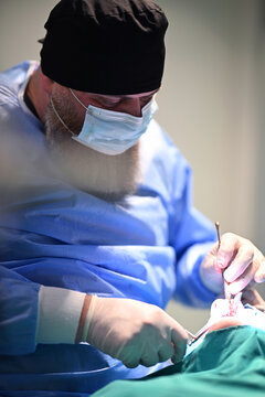 Bearded Surgeon Performing Oral Surgery In Operating Room Wearing Mask And Blue Scrubs